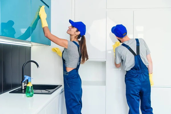 Пара профессиональных чистильщиков в синей униформе чистят мебель салфетками и брызгами на современной кухне . — стоковое фото
