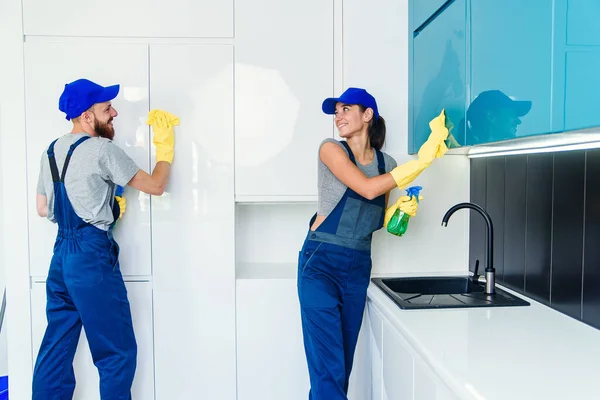 Paar professionele schoonmakers in blauw uniform reinigen het meubilair met doekjes en spray in de eigentijdse keuken. — Stockfoto