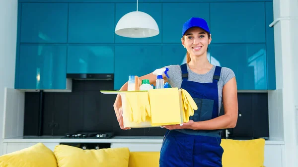 Smiling woman in special uniform for cleaning apartment holds the box with detergents, gloves, rags and standing in modern stylish kitchen. — Stock Photo, Image