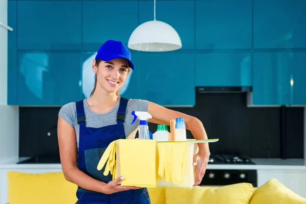 Улыбающаяся женщина в специальной униформе для уборки квартиры держит коробку с моющими средствами, перчатками, тряпками и стоя на современной стильной кухне . — стоковое фото