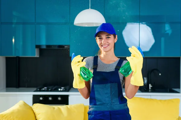 Молодая женщина в специальной одежде для чистки держит две бутылки различных моющих средств, стоя на высокотехнологичном кухонном фоне . — стоковое фото