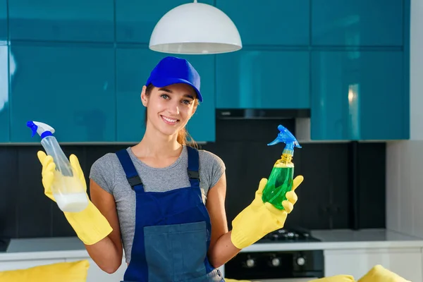 Θετική γυναίκα εργαζόμενος της επαγγελματικής υπηρεσίας καθαρισμού κάνει μια επιλογή μεταξύ των διαφόρων καθαριστικών παραγόντων στο παρασκήνιο της σύγχρονης κουζίνας. έννοια υπηρεσία καθαρισμού σπιτιού. — Φωτογραφία Αρχείου