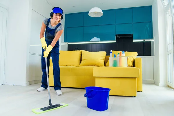 Mooie jonge vrouwelijke professionele schoonmaakster in speciaal uniform met hoofdtelefoon wassen van de vloer met dweil en luistert naar muziek in appartement. — Stockfoto