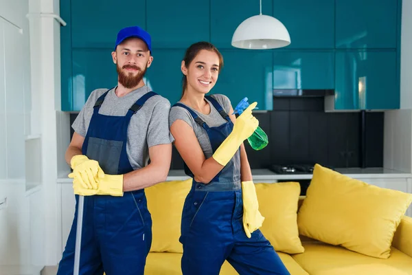 Aantrekkelijke man en mooie vrouw in workwears poseren op de camera in de hedendaagse keuken met detergenten — Stockfoto