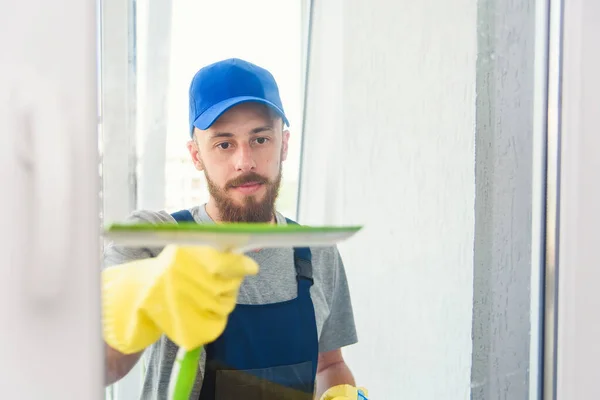Мужчина-уборщик, использующий скребок для мытья окна в офисе в фартуке и перчатках во время работы — стоковое фото