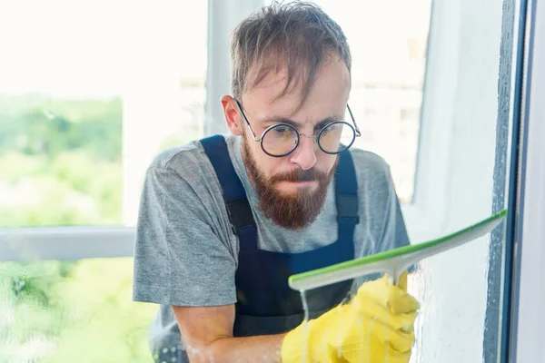Человек как профессиональный уборщик в синей униформе моющее окно с резиновой щеткой в помещении — стоковое фото