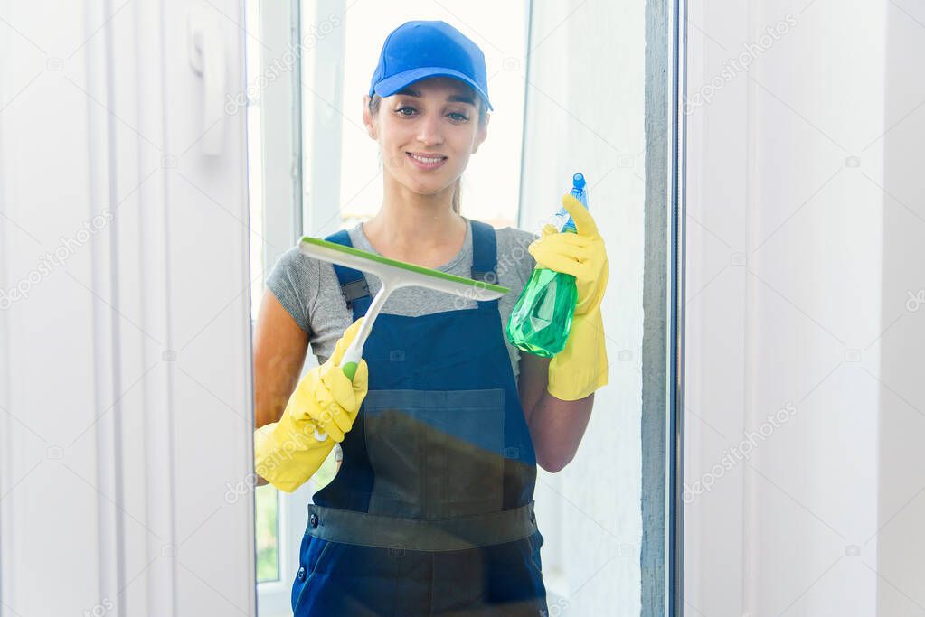 Professional worker from cleaning company washes window with a squeegee and detergents in modern apartment wearing special uniforms and yellow rubber gloves.