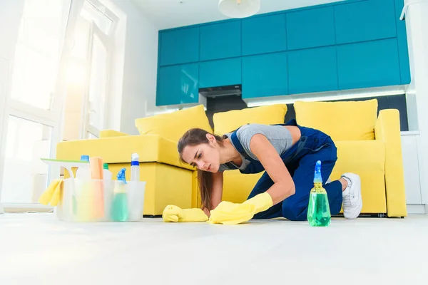 Jeune femme vêtue de vêtements spéciaux se tient sur ses genoux et le nettoyage du parquet en bois avec des détergents et chiffon sur la cuisine moderne . — Photo