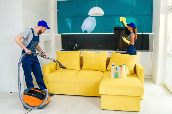 Young man with vacuum cleaner washing with vapor yellow sofa and beautiful woman wipes cuisine furniture.