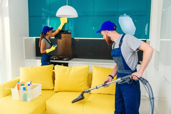 Young man vacuums yellow sofa and cute girl wiping kitchen furniture. Professional cleaners in apartment. — Stock Photo, Image