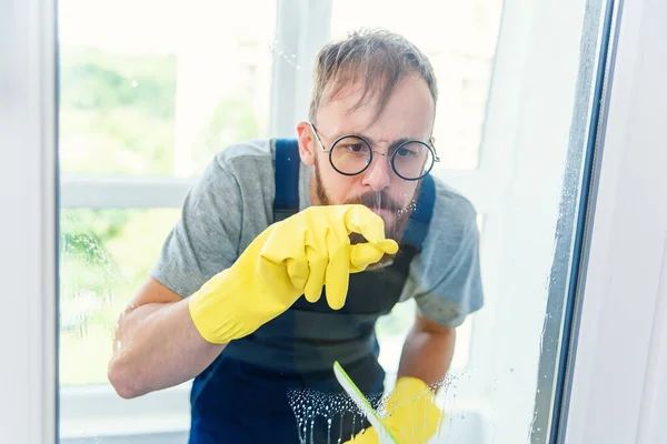 Homme barbu gai dans des lunettes drôles nettoie la fenêtre avec des détergents et enlever la saleté avec son doigt dans des gants . — Photo