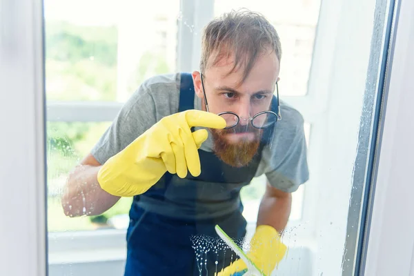 Homme barbu gai dans des lunettes drôles nettoie la fenêtre avec des détergents et enlever la saleté avec son doigt dans des gants . — Photo
