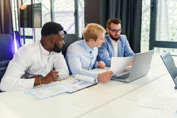 Tres hombres de negocios guapos de razas mixtas discutiendo oportunidades de su estrategia de negocio usando diferentes informes con infografías . — Foto de Stock