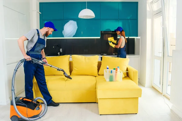 Young man vacuums yellow sofa and cute girl wiping kitchen furniture. Professional cleaners in apartment.