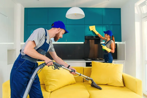 Jeune homme aspire canapé jaune et jolie fille essuyant meubles de cuisine. Nettoyeurs professionnels dans l'appartement . — Photo