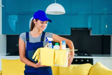 Bir kutu temizlik aletini elinde tutan kadın pembe arka planda yalnız başına kameraya bakıyor.