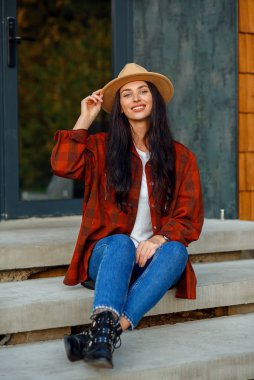 Kırmızı gömlekli, kot pantolonlu ve şapkalı güzel bir kadın ormandaki modern evinin merdivenlerinde oturuyor..