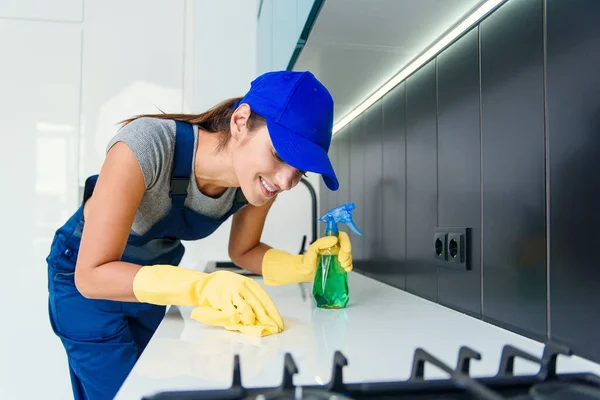 Jonge mooie vrouw in speciale uniform schoonmaken keukenmeubels met doek en wasmiddel. — Stockfoto