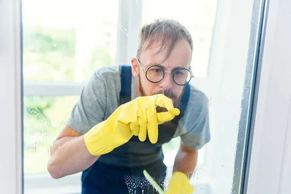 Веселый бородатый мужчина в смешных очках моет окно моющими средствами и удаляет грязь пальцем в перчатках . — стоковое фото