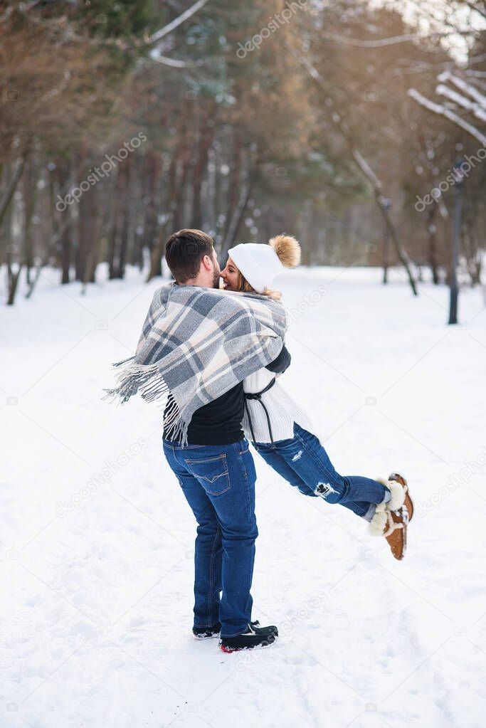 Young stylish couple in love have fun while walking on the snowy winter park. Handsome young man holds his beautiful girl on the hands.