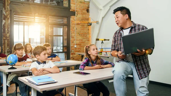 El profesor asiático se sienta en el escritorio con la computadora portátil en las manos y explica la lección para seis alumnos de la escuela primaria. Escuela Niños sentados en escritorios escuchando a su profesor . — Foto de Stock