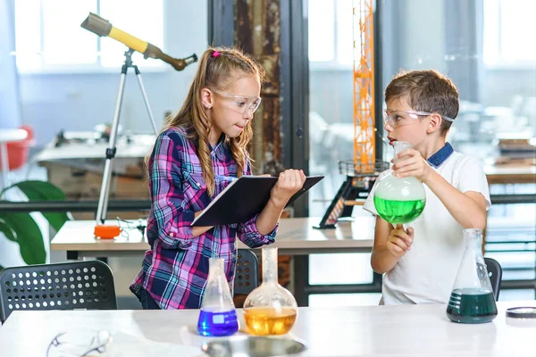 Children record results of the experiments in notebook. Two young clever caucasian pupils in protective glasses doing experiment with green liquid in beaker and dry ice.