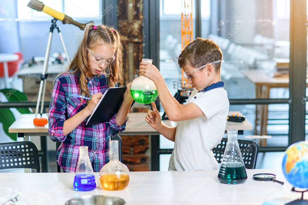 Children record results of the experiments in notebook. Two young clever caucasian pupils in protective glasses doing experiment with green liquid in beaker and dry ice.