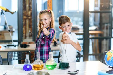 İki neşeli Kafkas bilim adamı çocuğunun güvenlik gözlüğüyle ilkokul kimya dersinde başparmaklarını gösteren portre fotoğrafı. Bilim, kimya ve çocuklar. Yavaş çekim