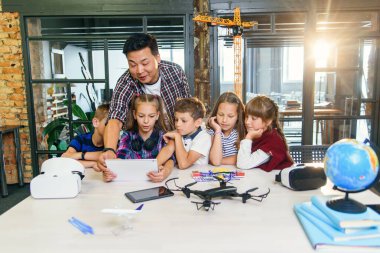 Asyalı öğretmen teknoloji dersinde dijital cihazlar kullanan beş genç öğrenciyle çalışıyor. Eğitim, bilim, gelişme ve modern teknoloji kavramı.