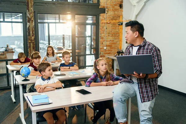 El profesor asiático se sienta en el escritorio con la computadora portátil en las manos y explica la lección para seis alumnos de la escuela primaria. Escuela Niños sentados en escritorios escuchando a su profesor . — Foto de Stock