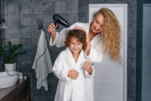 Beautiful mom dries hair with a hairdryer to her little happy son, dressed in a white bathrobe. — Stock Photo, Image