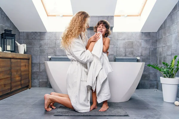 Atractiva madre joven con pelo rizado claro en bata de baño arrodillándose y limpiando con toalla blanca a su pequeño hijo guapo . — Foto de Stock