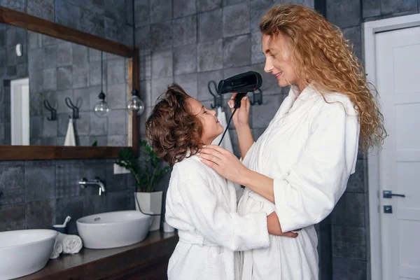 Beautiful mom dries hair with a hairdryer to her little happy son, dressed in a white bathrobe. — Stock Photo, Image