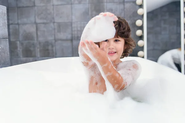 Vtipný chlapeček si hraje s pěnou ve vaně. — Stock fotografie