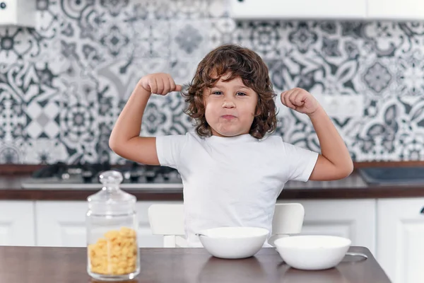 Happy little boy showing biceps muscles while eating healthy breakfast of milk and cornflakes at the stylish kitchen in the morning.