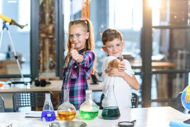 İki neşeli Kafkas bilim adamı çocuğunun güvenlik gözlüğüyle ilkokul kimya dersinde başparmaklarını gösteren portre fotoğrafı. Bilim, kimya ve çocuklar. Yavaş çekim