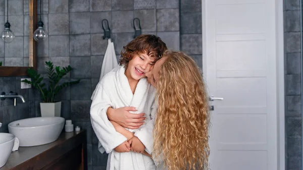 Beautiful mother with curly hair in a white bathrobe hugs her little smiling son in the bathroom. — Stock Photo, Image