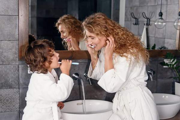 Atractiva y sonriente madre joven en bata limpia sus dientes junto con su pequeño hijo guapo cerca del lavabo con espejo. Concepto de familia feliz . — Foto de Stock