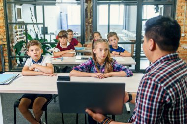 Genç Koreli erkek öğretmen dizüstü bilgisayarla masada oturuyor ve altı ilkokul öğrencisine ders veriyor. Okul çocukları sıralarında oturup konferansı dinliyorlar..