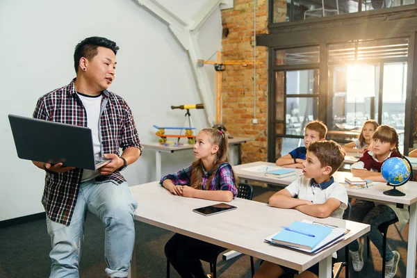 Joven profesor coreano sentado en el escritorio con portátil dando lección para seis alumnos de la escuela primaria. Escuela Los niños sentados en los escritorios y escuchar la conferencia . — Foto de Stock