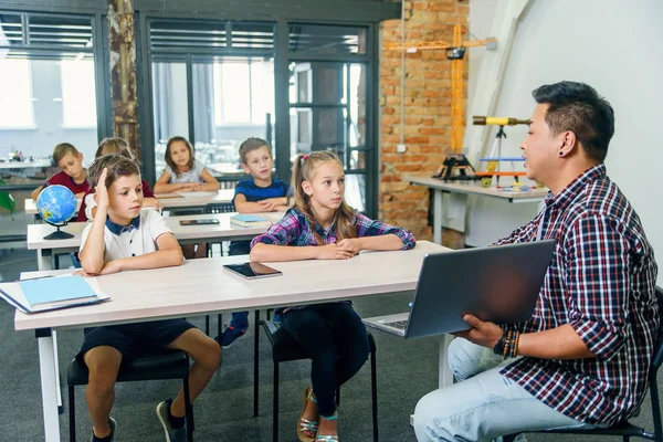 El chico de la escuela se sienta detrás del escritorio junto con sus compañeros de clase y levanta la mano para hacer preguntas a su maestro durante la lección en la escuela primaria. . —  Fotos de Stock