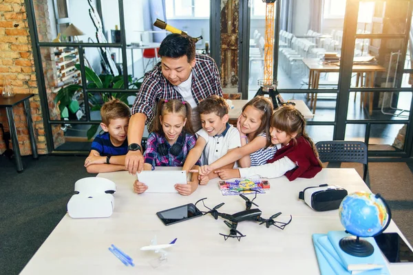 School teacher at desk works with five young pupils using digital tablet computer in technology class. Development, technology and modern education concept.