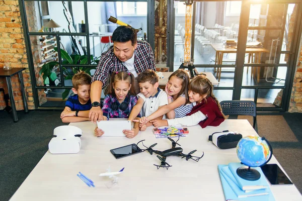 School teacher at desk works with five young pupils using digital tablet computer in technology class. Development, technology and modern education concept.