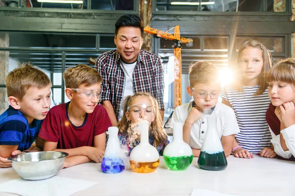 Asyalı öğretmen modern okuldaki çocuklar için kuru buzla deneyler yapıyor. Deney sırasında bilim adamı reaksiyon dumanı ve renkli sıvıyı gösteren bir matara tutuyor.. — Stok fotoğraf