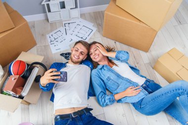 Yeni evli çift, yeni dairelerinin ahşap zemininde uzanmış akıllı telefonlarıyla selfie çekiyorlar. Üst görünüm.