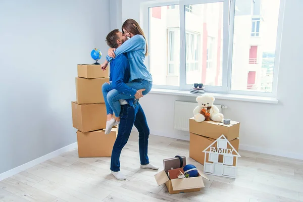 Front view of pretty smiling girl which holds by her handsome young boyfriend in the middle of their new flat full of cardboard boxes. — Stock Photo, Image