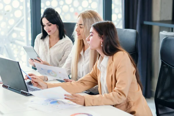 Jovenes trabajadoras de oficina sentadas en la sala de reuniones y discutiendo su proyecto de negocio conjunto usando datos financieros y computadoras . — Foto de Stock