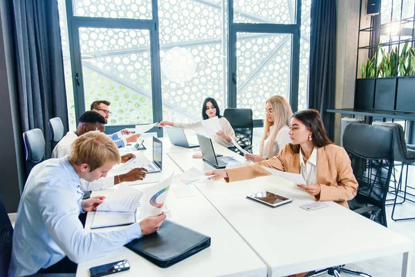 Compañeros de carreras mixtas analizando diferentes papeles de negocios y trabajando con computadoras en las mesas de la oficina moderna . — Foto de Stock