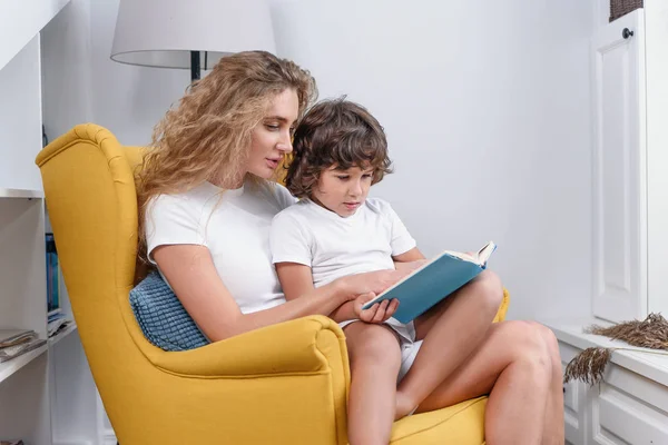 Κοντινό πλάνο της ελκυστική νεαρή συμπυκνωμένη μαμά η οποία κάθεται σε μαλακό πράσινο καρέκλα με 5-γερασμένη χαρούμενη γιο της και απολαμβάνοντας την ανάγνωση ενδιαφέρον βιβλίο — Φωτογραφία Αρχείου