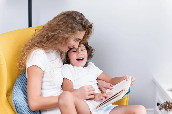 Belo retrato de sorrir feliz mãe e filho em roupas brancas que se divertem juntos enquanto lêem um livro interessante — Fotografia de Stock